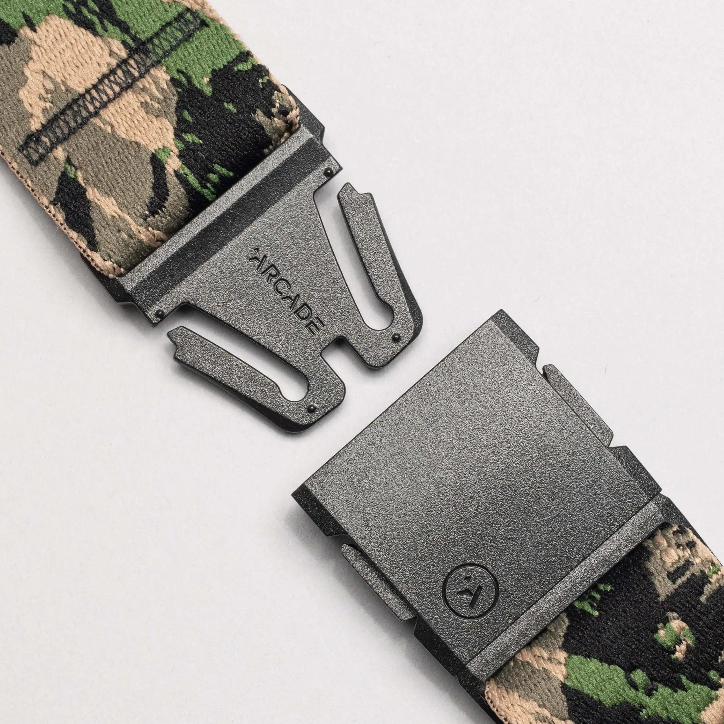 A close-up of an Arcade Belts Island Collection travel belt with camouflage stretch webbing and a black plastic buckle, partially unclasped. The clasp is engraved with ARCADE against a light, neutral background.