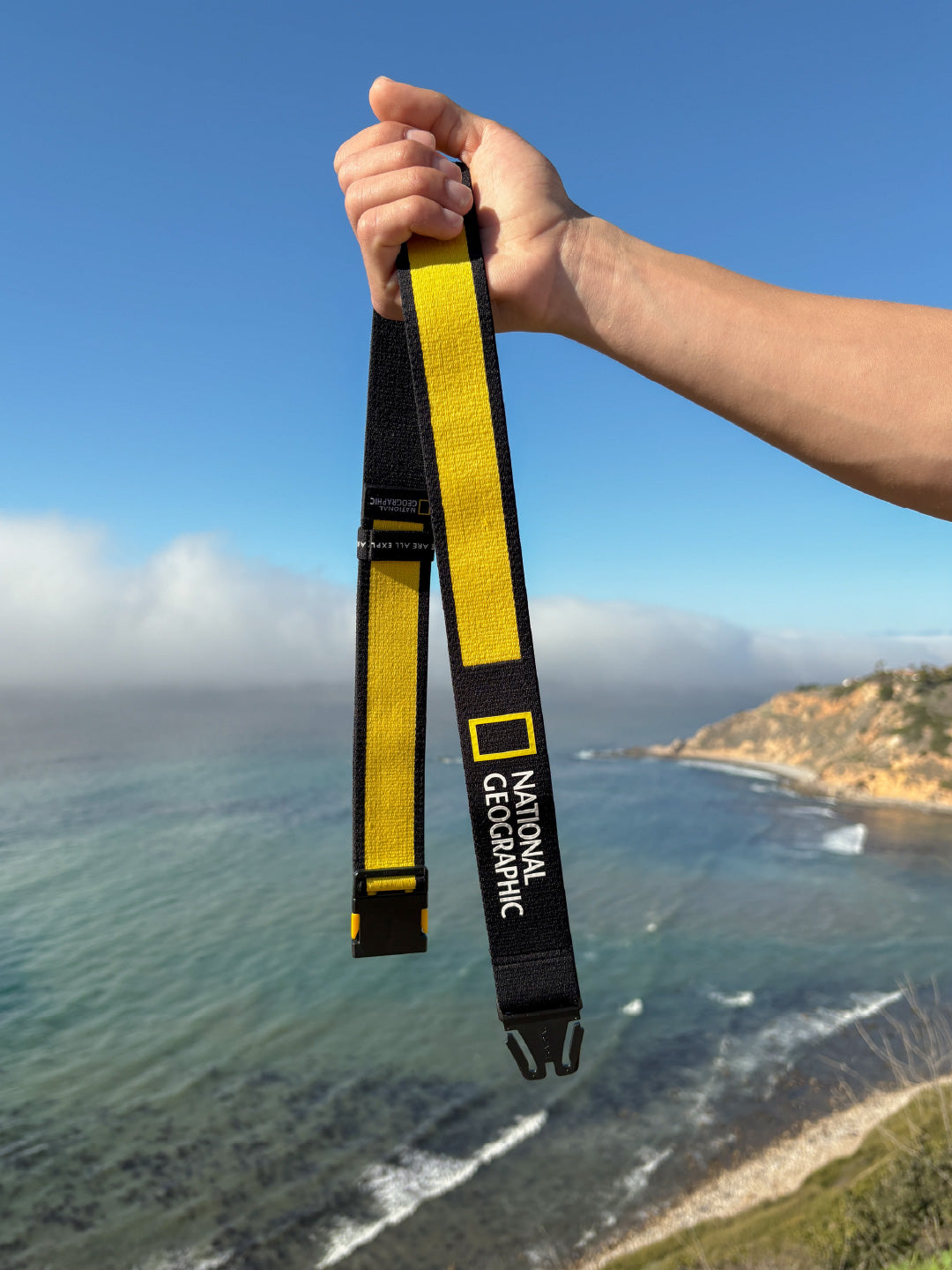 A hand holds up a black and yellow National Geographic-branded strap against a coastal backdrop, with cliffs, ocean waves, and a partly cloudy sky in the background.