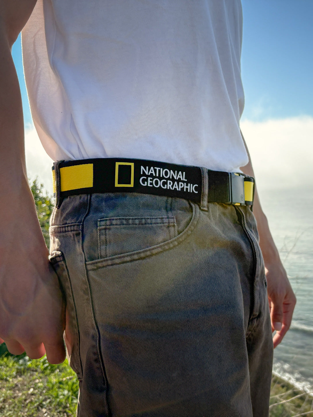 A person wearing faded jeans and a white shirt stands outdoors near water, with a visible black and yellow National Geographic belt around their waist. The background shows greenery and a hazy sky.