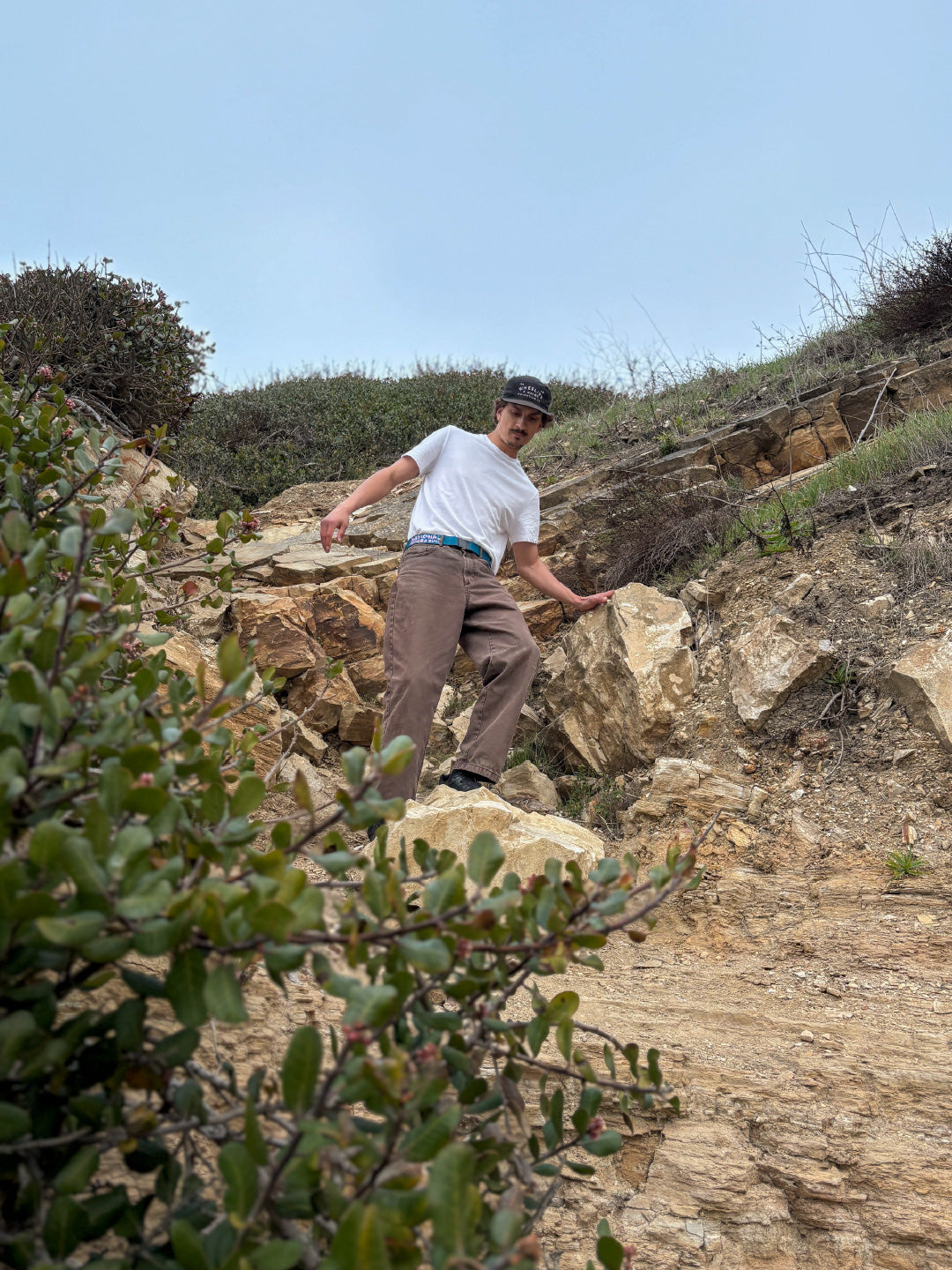 A person wearing a white T-shirt, brown pants, and a cap stands on a rocky, sloped hillside surrounded by shrubs and greenery under a cloudy sky.