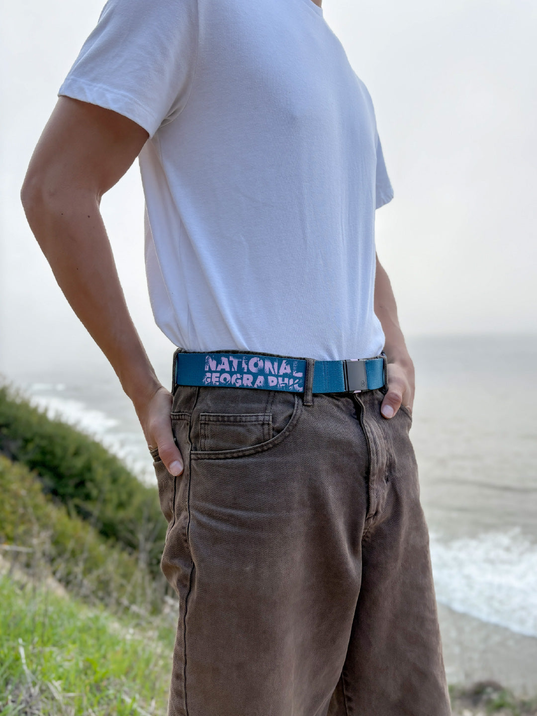 A person stands outdoors by the ocean, wearing a white t-shirt and brown pants. Their belt is blue and says NATIONAL GEOGRADER. The background includes grassy cliffs and the sea under a cloudy sky.