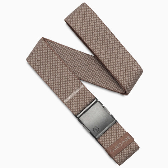 The Arcade Belts Carry is a taupe textured fabric belt with performance stretch webbing, a black rectangular buckle, and a small leather ARCADE tag near the end. It’s displayed neatly folded in a loose zigzag pattern.