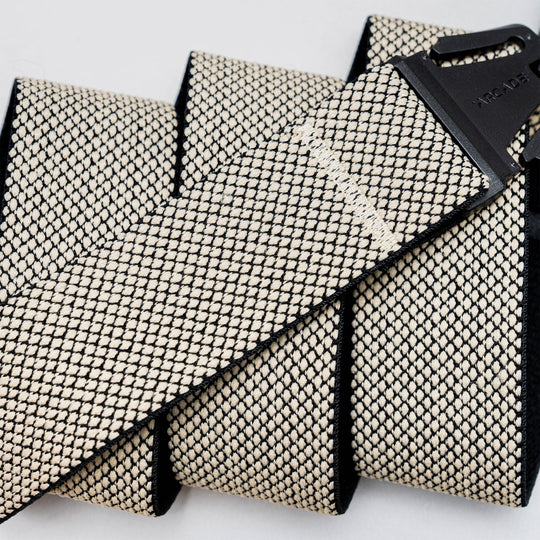 A close-up of the Arcade Belts Carry strap in beige and black, featuring a unique weave with an A2 buckle, shown coiled and laid out on a light background.