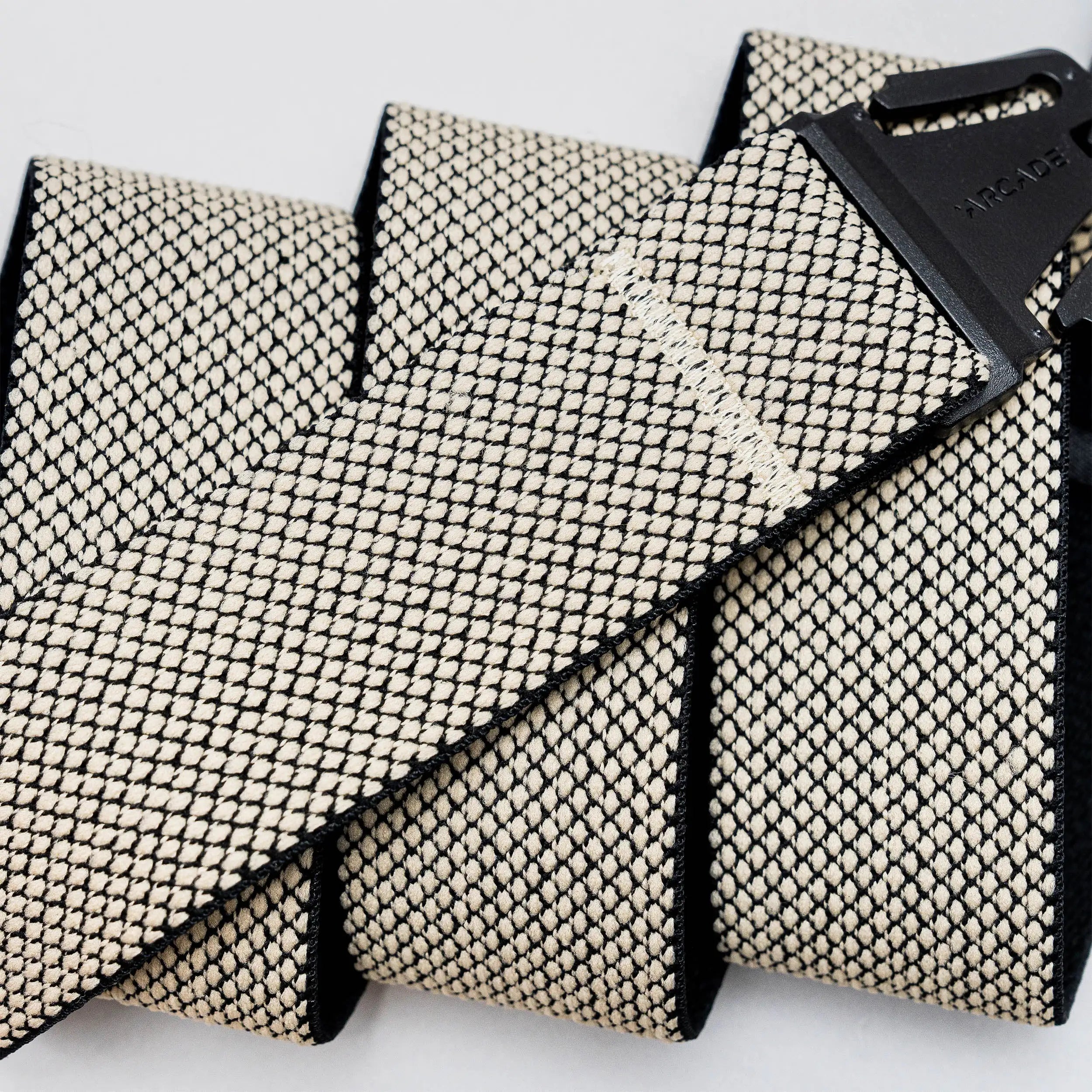 A close-up of the Arcade Belts Carry strap in beige and black, featuring a unique weave with an A2 buckle, shown coiled and laid out on a light background.