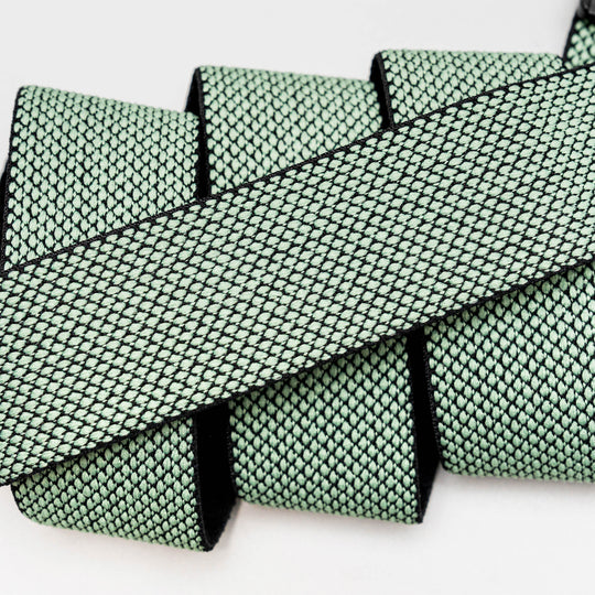 A close-up of the Arcade Belts Carry strap, featuring light green woven fabric with a black diamond pattern, highlights its unique weave in overlapping layers against a neutral background.