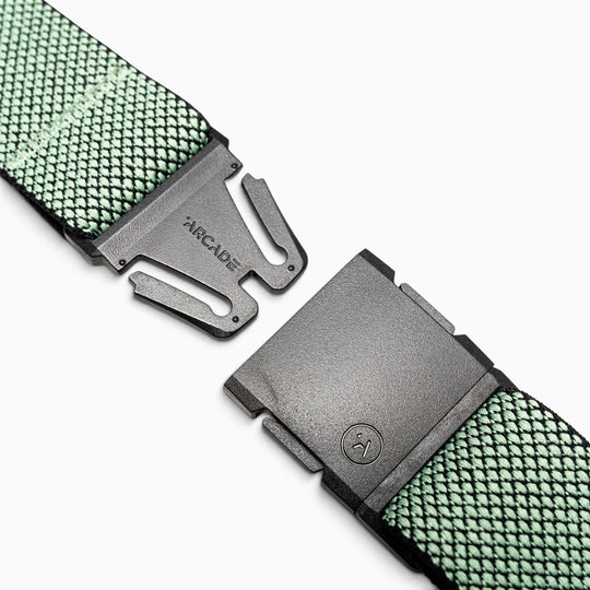 A close-up of the green Arcade Belts Carry woven fabric belt with a black plastic A2 buckle, shown unclasped on a white background. The buckle features ARCADE branding, while the unique weave and performance stretch webbing ensure exceptional comfort and flexibility.