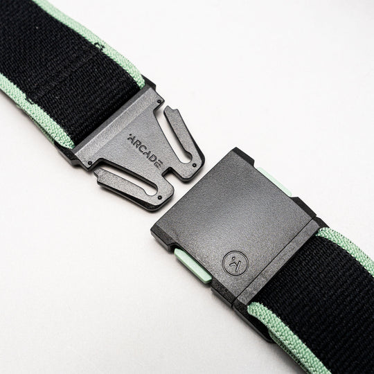 A close-up of the Arcade Belts Carto in black and mint green, featuring a black plastic buckle partially unclasped, with the Arcade Belts name visible on the metal prong section against a light background.