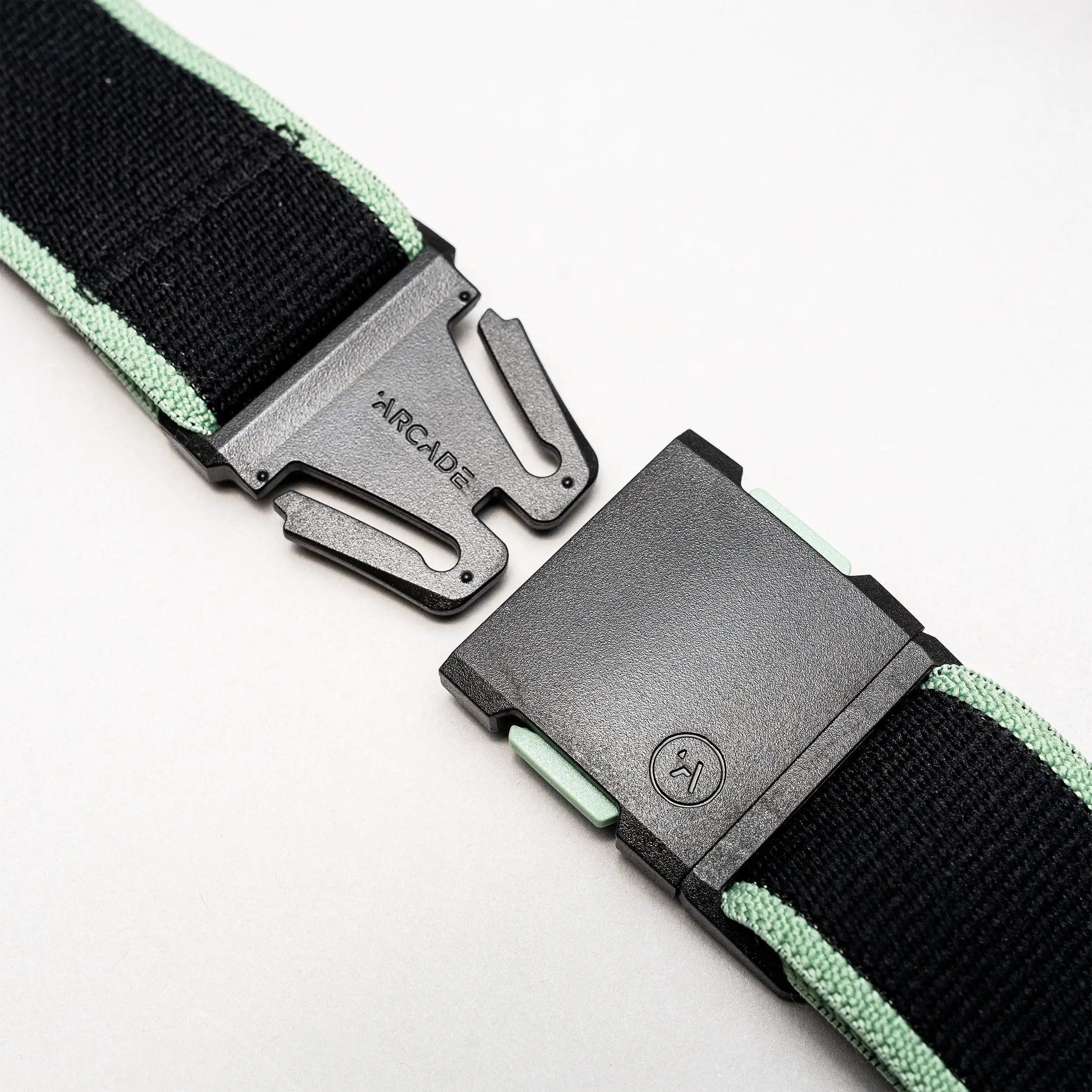 A close-up of the Arcade Belts Carto in black and mint green, featuring a black plastic buckle partially unclasped, with the Arcade Belts name visible on the metal prong section against a light background.