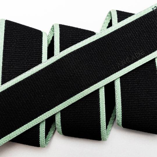 A close-up of the Arcade Belts Carto, a black stretch belt with light green edges, folded in a zigzag on a white background. The word ARCADE is subtly embossed on the webbing.
