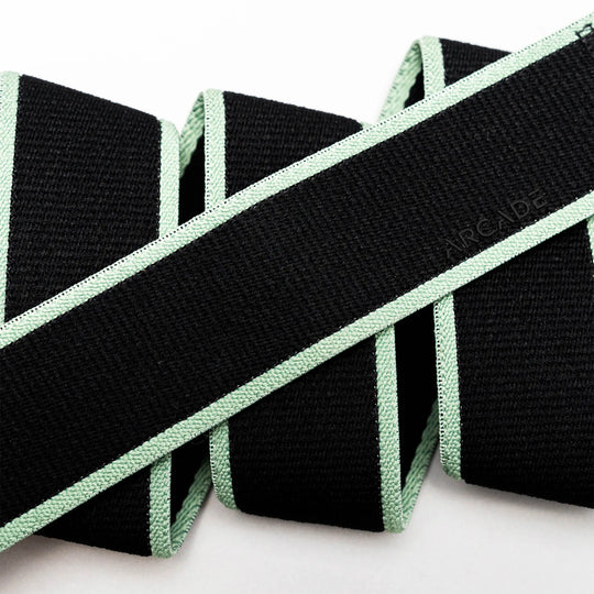 A close-up of the Arcade Belts Carto, a black stretch belt with light green edges, folded in a zigzag on a white background. The word ARCADE is subtly embossed on the webbing.