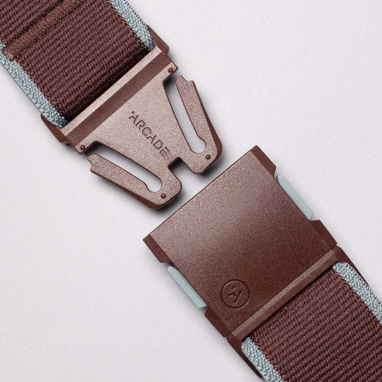 Close-up of the Arcade Belts Carto, a low-profile elastic stretch belt with a plastic snap buckle. One side is labeled Arcade, with light blue edge stitching, set against a light gray background.