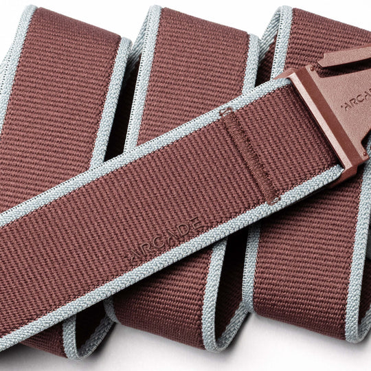 A close-up image of the Arcade Belts Carto, a brown woven stretch webbing belt with light blue edging and a plastic buckle. The ARCADE logo is embossed near the buckle, showcasing its quality craftsmanship for travel use.