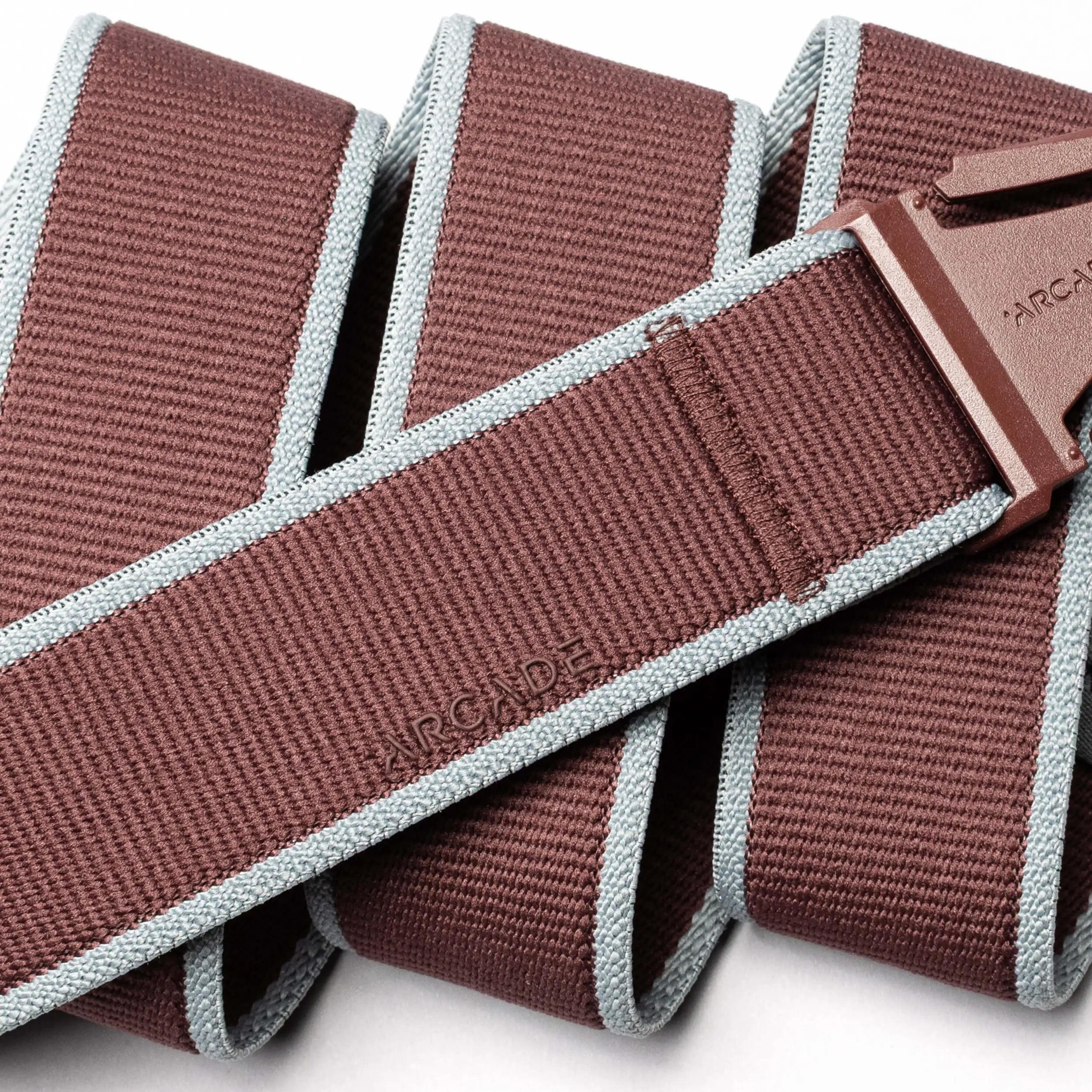 A close-up image of the Arcade Belts Carto, a brown woven stretch webbing belt with light blue edging and a plastic buckle. The ARCADE logo is embossed near the buckle, showcasing its quality craftsmanship for travel use.