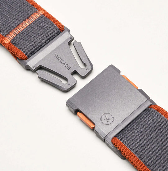 Close-up of a grey and orange Carto belt by Arcade Belts, made from nylon elastic stretch material with a metal magnetic buckle featuring the brand name and logo, partially fastened on a white background.