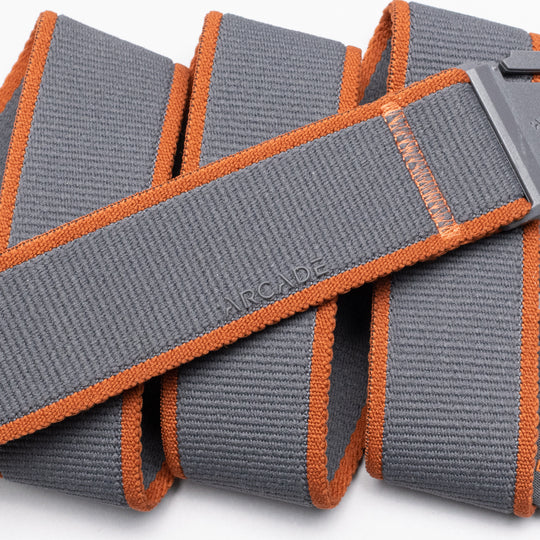 A close-up of the Carto, a gray elastic stretch belt by Arcade Belts, featuring orange edges and a rectangular plastic buckle. ARCADE is embossed on the coiled belt, highlighting its texture and stitching—perfect for travel belt lovers.