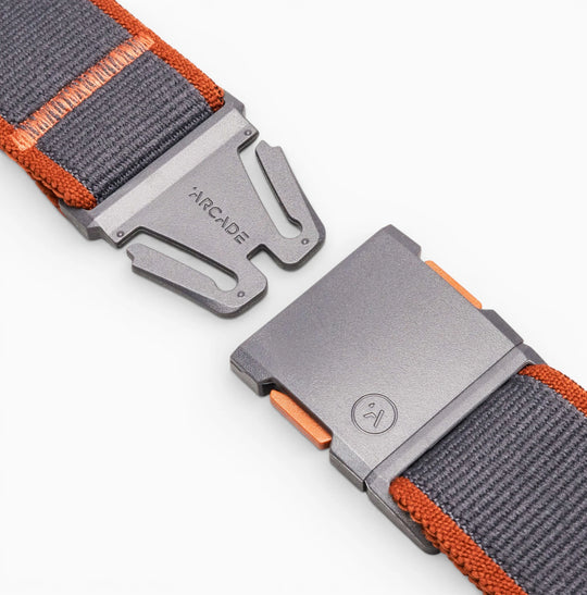 Close-up of a grey and orange Carto belt by Arcade Belts, made from nylon elastic stretch material with a metal magnetic buckle featuring the brand name and logo, partially fastened on a white background.