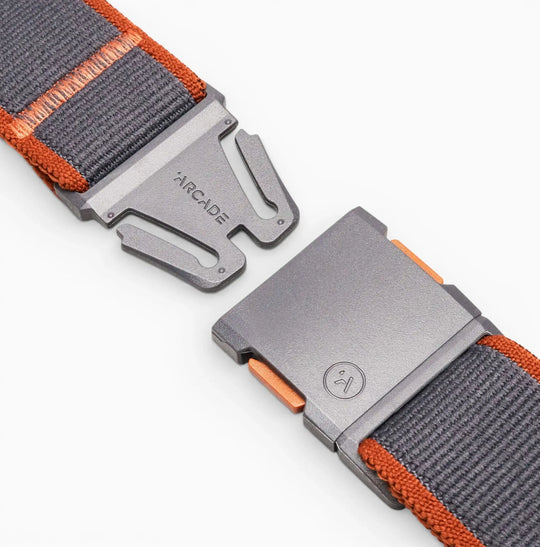 Close-up of a grey and orange Carto belt by Arcade Belts, made from nylon elastic stretch material with a metal magnetic buckle featuring the brand name and logo, partially fastened on a white background.