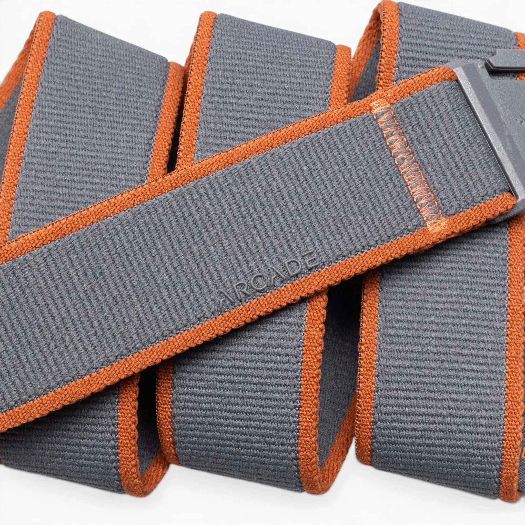 A close-up of the Carto, a gray elastic stretch belt by Arcade Belts, featuring orange edges and a rectangular plastic buckle. ARCADE is embossed on the coiled belt, highlighting its texture and stitching—perfect for travel belt lovers.