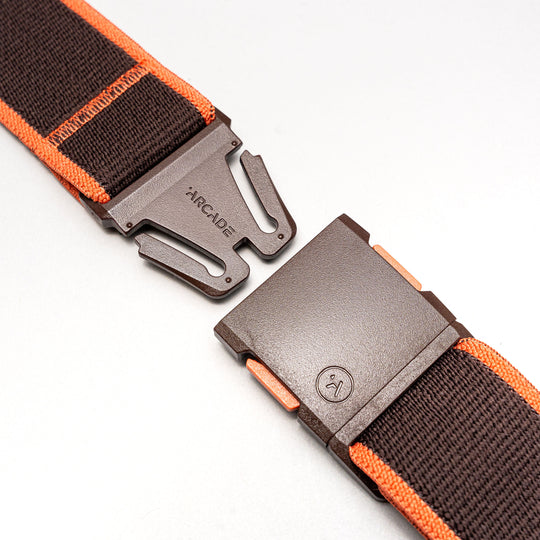 A close-up of the Arcade Belts Carto elastic stretch belt in dark brown with orange edges, featuring a matte brown metal buckle engraved with ARCADE, displayed open on a light gray surface.