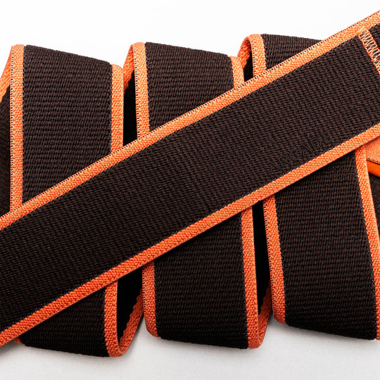 A close-up of Carto by Arcade Belts reveals brown textured straps with bright orange edges, overlapping on a white background to highlight the versatility of their stretch webbing.