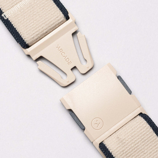 A close-up of the Carto belt by Arcade Belts shows a partially unclasped beige plastic buckle engraved with ARCADE, attached to a navy and beige striped woven strap, highlighting the brands signature low-profile design.