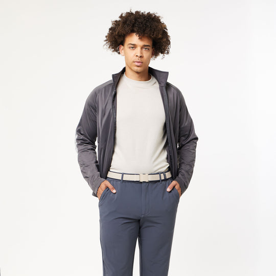 A young man with curly hair, wearing a light gray t-shirt, dark gray zip-up jacket, and blue-gray pants with the Arcade Belts Carto Kit featuring a low-profile plastic buckle, stands with hands in his pockets against a plain white background.