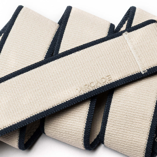 A close-up of the Carto belt by Arcade Belts, featuring a beige color with dark blue edges, a low-profile design, and ARCADE embossed on the surface. The belt is shown coiled loosely.
