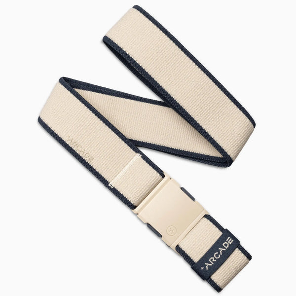 The Arcade Belts Carto is a beige stretch webbing travel belt with black edges and a matching beige plastic buckle, ARCADE printed near the end. It is loosely coiled on a white background.