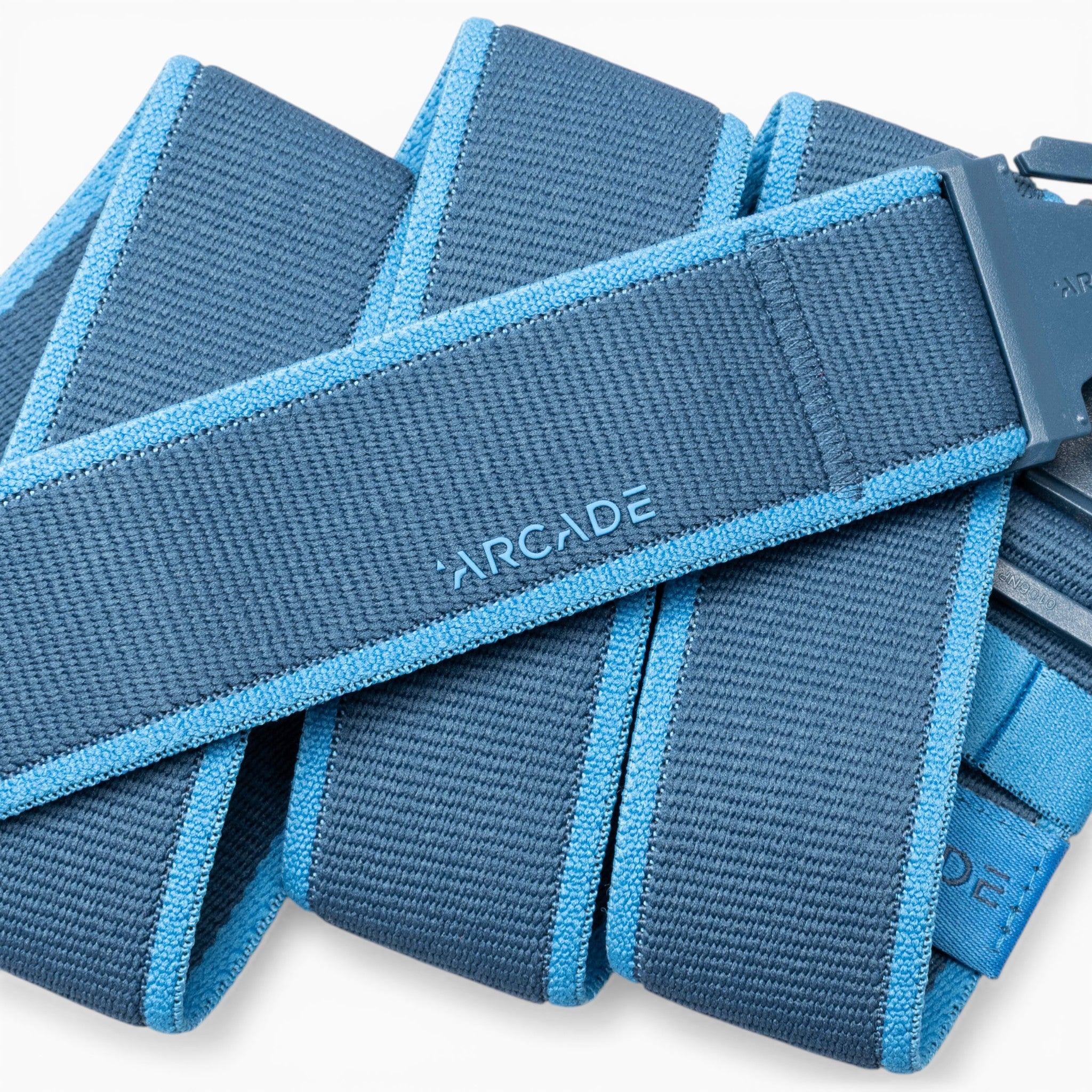 A Carto belt by Arcade Belts, in blue with light blue edges, features a woven performance stretch design. Coiled in several layers and equipped with a plastic buckle embossed with “ARCADE” on the strap, it’s the ideal accessory for adventure seekers who prioritize both comfort and style.