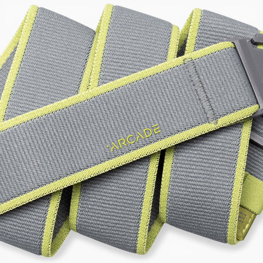 A close-up of the Arcade Belts Carto: a coiled gray performance stretch belt featuring light green edges and “ARCADE” stitched in yellow, with part of a gray buckle visible—ideal for adventure.