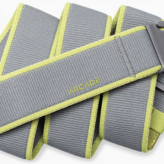A close-up of the Arcade Belts Carto: a coiled gray performance stretch belt featuring light green edges and “ARCADE” stitched in yellow, with part of a gray buckle visible—ideal for adventure.