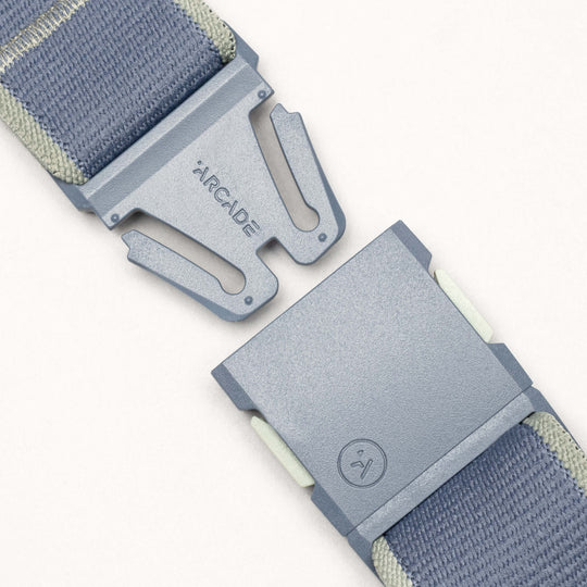 A close-up of the Arcade Belts Carto, a blue and gray performance stretch belt with a partially unfastened metal buckle engraved with ARCADE and a small logo, set against a light background.