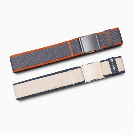 The Carto Kit by Arcade Belts includes two elastic travel belts with plastic buckles—one dark gray with orange edges, the other beige with dark blue edges—each featuring the ARCADE label. Shown on a light background.