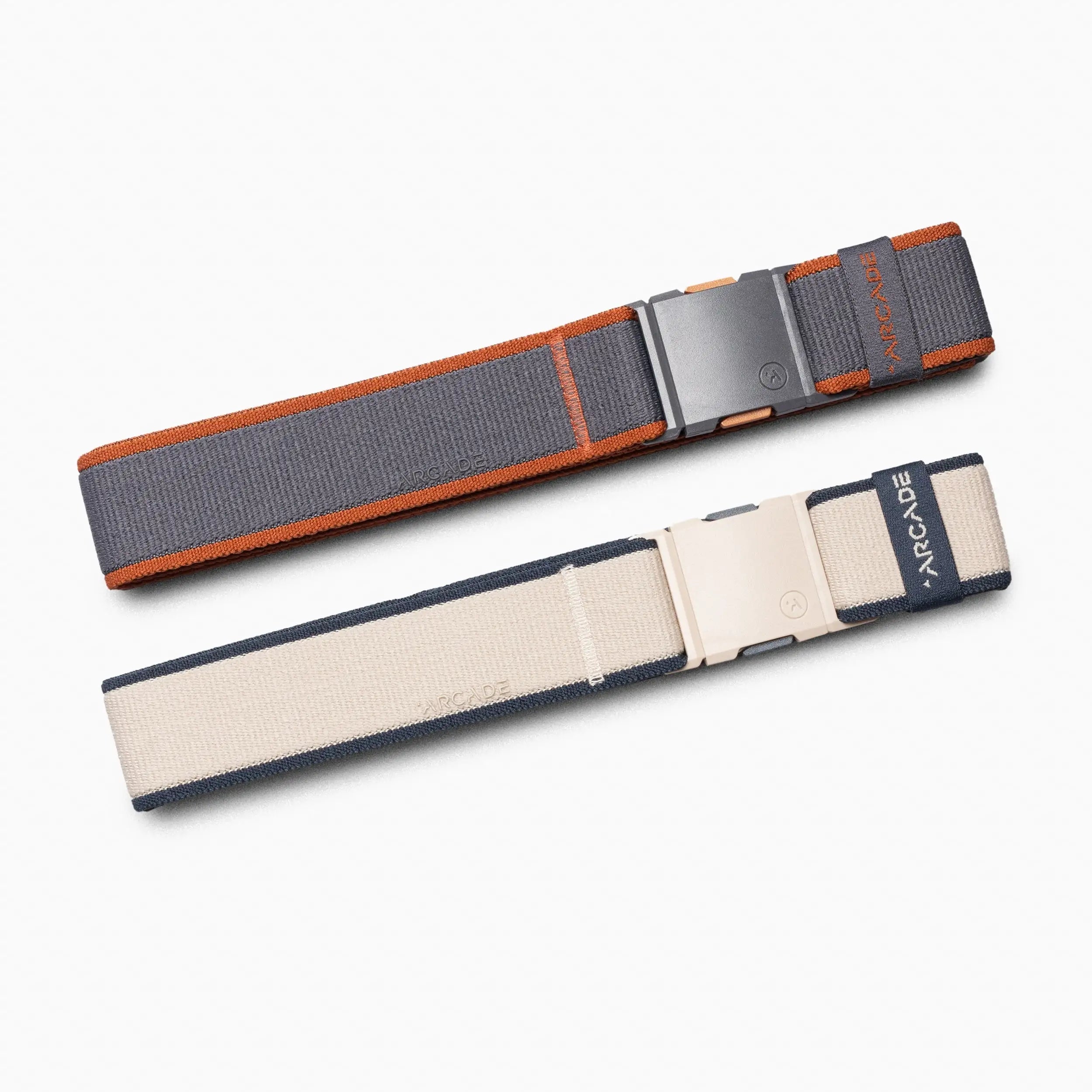The Carto Kit by Arcade Belts includes two elastic travel belts with plastic buckles—one dark gray with orange edges, the other beige with dark blue edges—each featuring the ARCADE label. Shown on a light background.