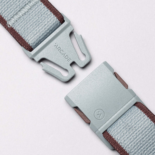 Close-up of a light gray plastic buckle on the Carto Elastic Stretch Belt by Arcade Belts, featuring brown edging, partially unfastened against a light background. The word ARCADE is printed on one side, reflecting the brands signature style.