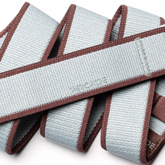 A close-up of the Arcade Belts Carto, a light gray stretch webbing belt with brown edges, coiled in overlapping loops. The word ARCADE is subtly embossed, showcasing the brand’s signature style.