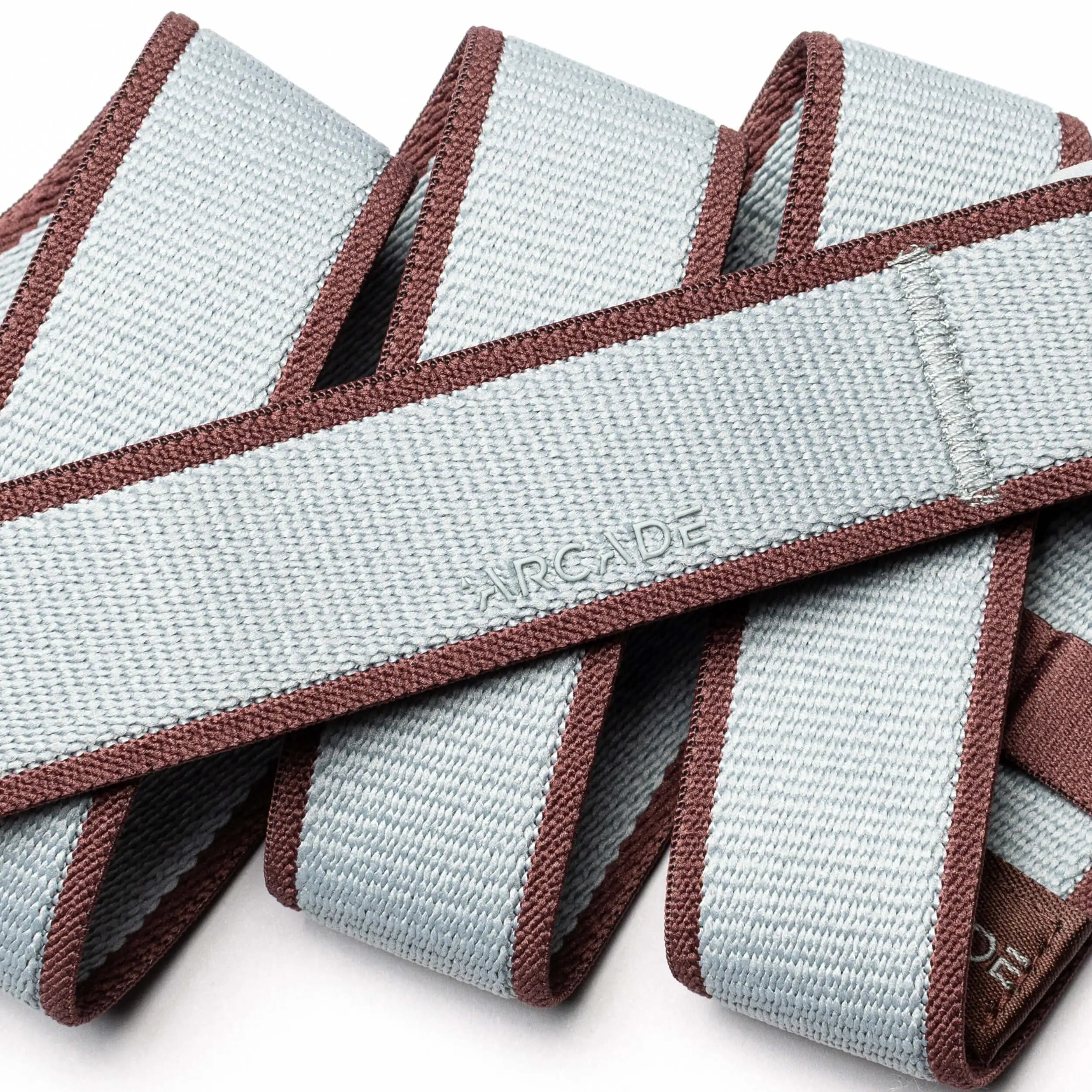 A close-up of the Arcade Belts Carto, a light gray stretch webbing belt with brown edges, coiled in overlapping loops. The word ARCADE is subtly embossed, showcasing the brand’s signature style.