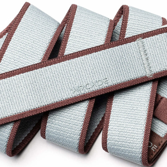 A close-up of the Arcade Belts Carto, a light gray stretch webbing belt with brown edges, coiled in overlapping loops. The word ARCADE is subtly embossed, showcasing the brand’s signature style.