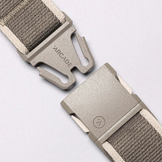 A close-up of the Arcade Belts Carto, a gray performance stretch belt with a plastic quick-release buckle, featuring the Arcade logo on one side and a small Carto smiley face logo on the other, laid on a light gray background.