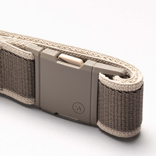 A close-up of the Arcade Belts Carto performance stretch belt in beige and gray with a rectangular plastic buckle, neatly rolled on a white surface—perfect for your next adventure.