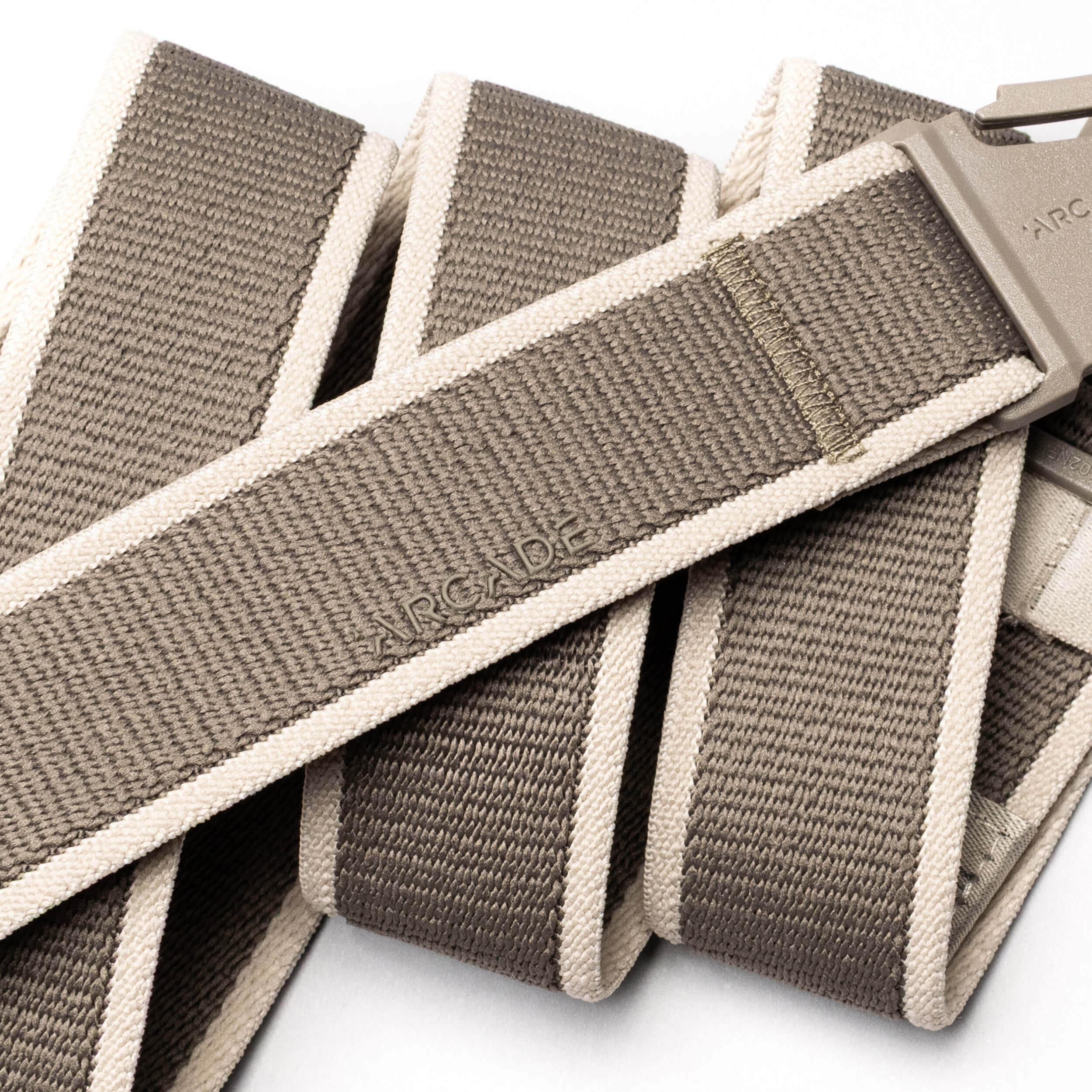 A close-up of the Carto belt by Arcade Belts in taupe and beige, featuring a plastic buckle and "ARCADE" embossed on the stretch fabric. The coiled performance belt, ideal for adventure, is shown against a white background.