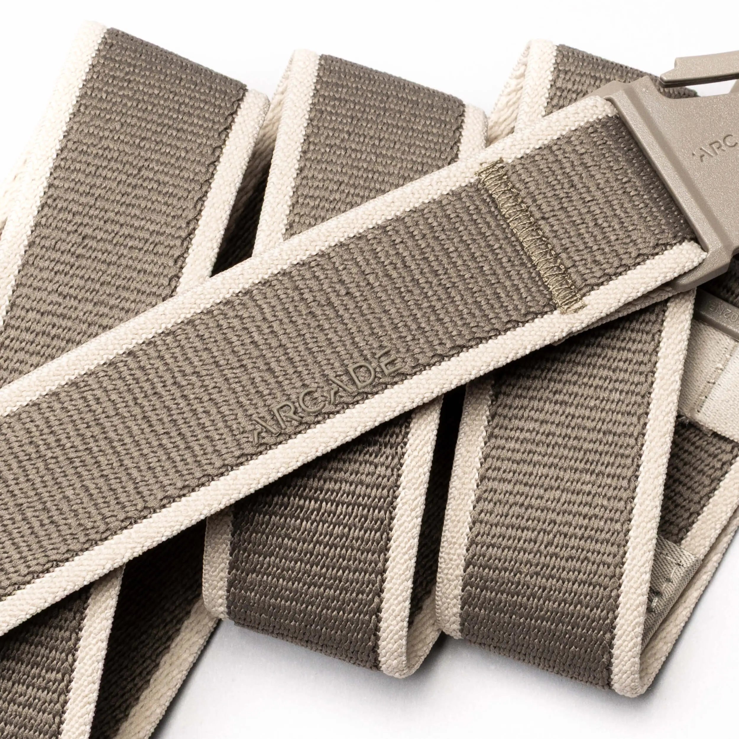 A close-up of the Carto belt by Arcade Belts in taupe and beige, featuring a plastic buckle and "ARCADE" embossed on the stretch fabric. The coiled performance belt, ideal for adventure, is shown against a white background.