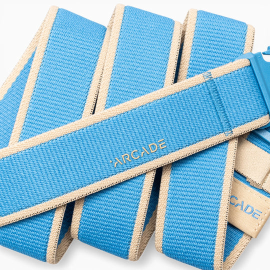 A close-up of the Arcade Belts Carto, a blue and beige woven performance stretch belt with “ARCADE” stitched in yellow on the fabric. Designed for adventure, this coiled belt features a plastic buckle and blends style with functionality for active lifestyles.