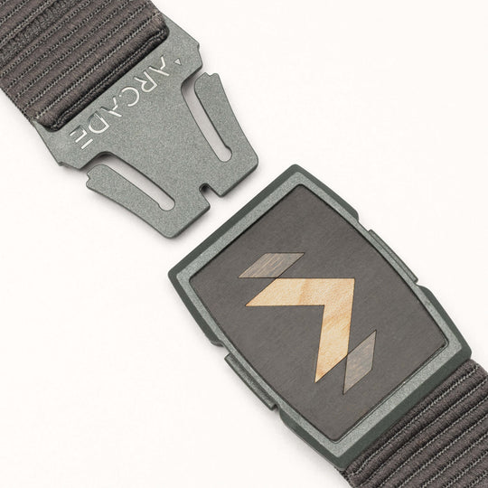 A close-up of the dark grey Carver stretch belt by Arcade Belts, featuring a partially unfastened metal buckle with geometric wood inlay and ARCADE engraved on the clasp, plus a textured strap.