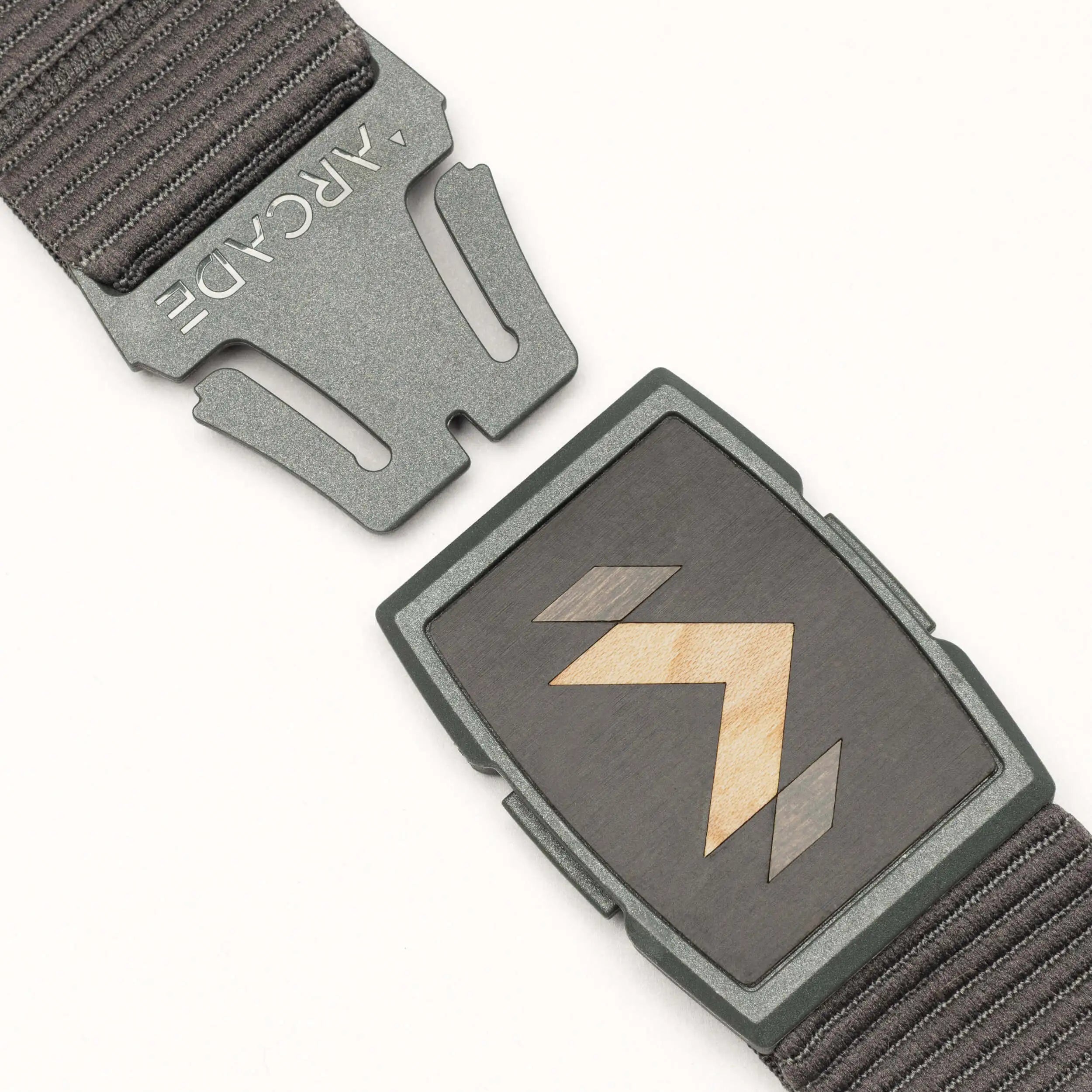 A close-up of the dark grey Carver stretch belt by Arcade Belts, featuring a partially unfastened metal buckle with geometric wood inlay and ARCADE engraved on the clasp, plus a textured strap.