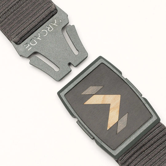 A close-up of the dark grey Carver stretch belt by Arcade Belts, featuring a partially unfastened metal buckle with geometric wood inlay and ARCADE engraved on the clasp, plus a textured strap.