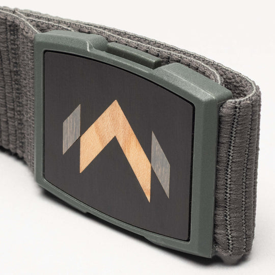 A close-up of the Arcade Belts Carver: a gray stretch belt with a rectangular buckle featuring a black face and geometric wood inlay with light and dark wood tones—a signature Carver detail.