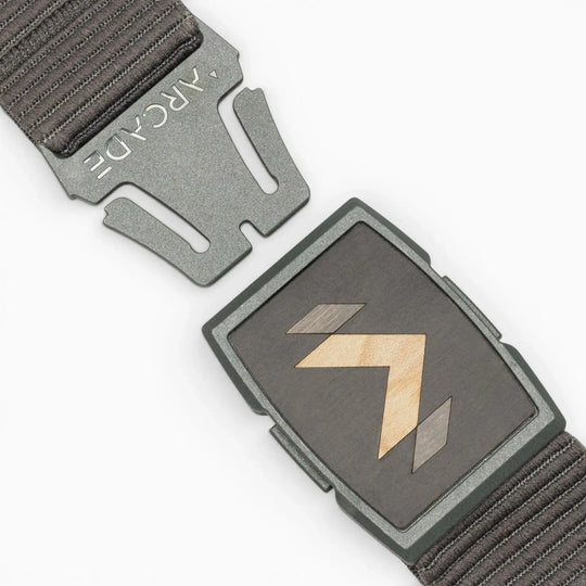 A close-up of the dark grey Carver stretch belt by Arcade Belts, featuring a partially unfastened metal buckle with geometric wood inlay and ARCADE engraved on the clasp, plus a textured strap.