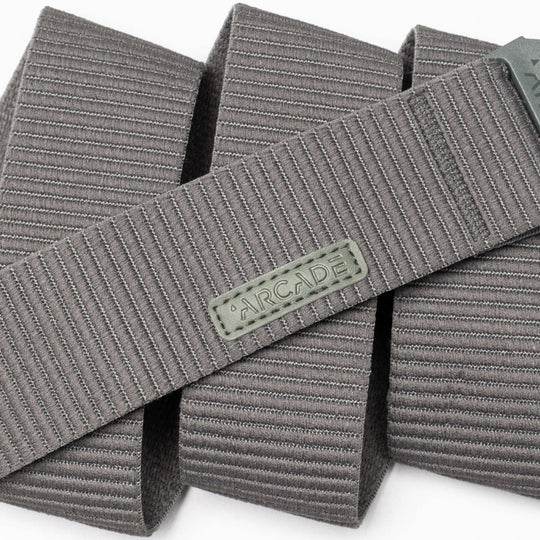 A close-up of the gray Carver stretch belt by Arcade Belts, featuring a ribbed texture and a rectangular Arcade logo patch, shown in overlapping folds on a white background.