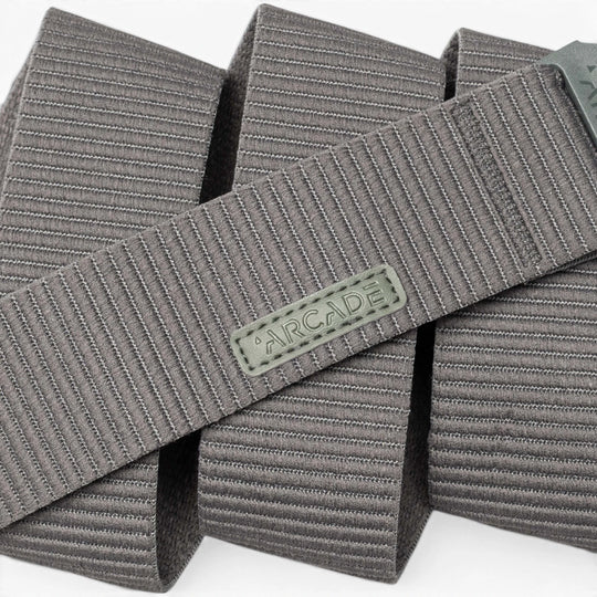 A close-up of the gray Carver stretch belt by Arcade Belts, featuring a ribbed texture and a rectangular Arcade logo patch, shown in overlapping folds on a white background.